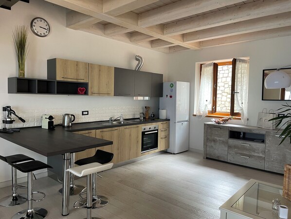 A modern kitchen with wooden cabinets, a built-in oven, and a refrigerator. There are two bar stools and a window with curtains.