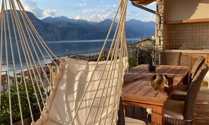 A hammock chair overlooking a lake with mountains in the background, a wooden table, wicker chairs, and a cat sitting on the table.