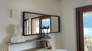 Ein Zimmer mit einem großen Spiegel, einer Buddha-Statue, einer Vase mit Trockenblumen und Meerblick durch ein Fenster.