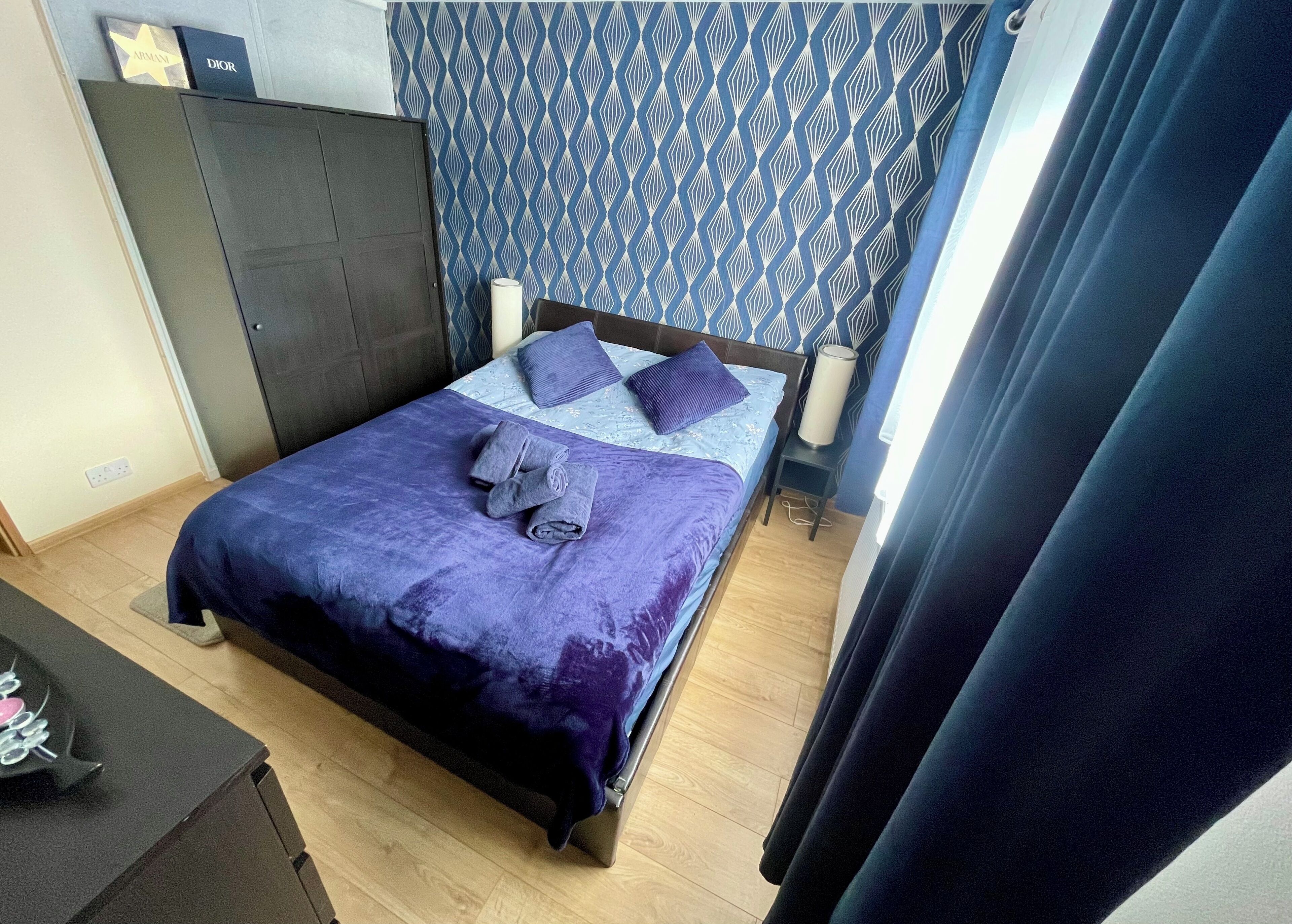 A neatly made bed with a purple blanket and pillows, a wooden floor, a wardrobe, and a patterned wall.