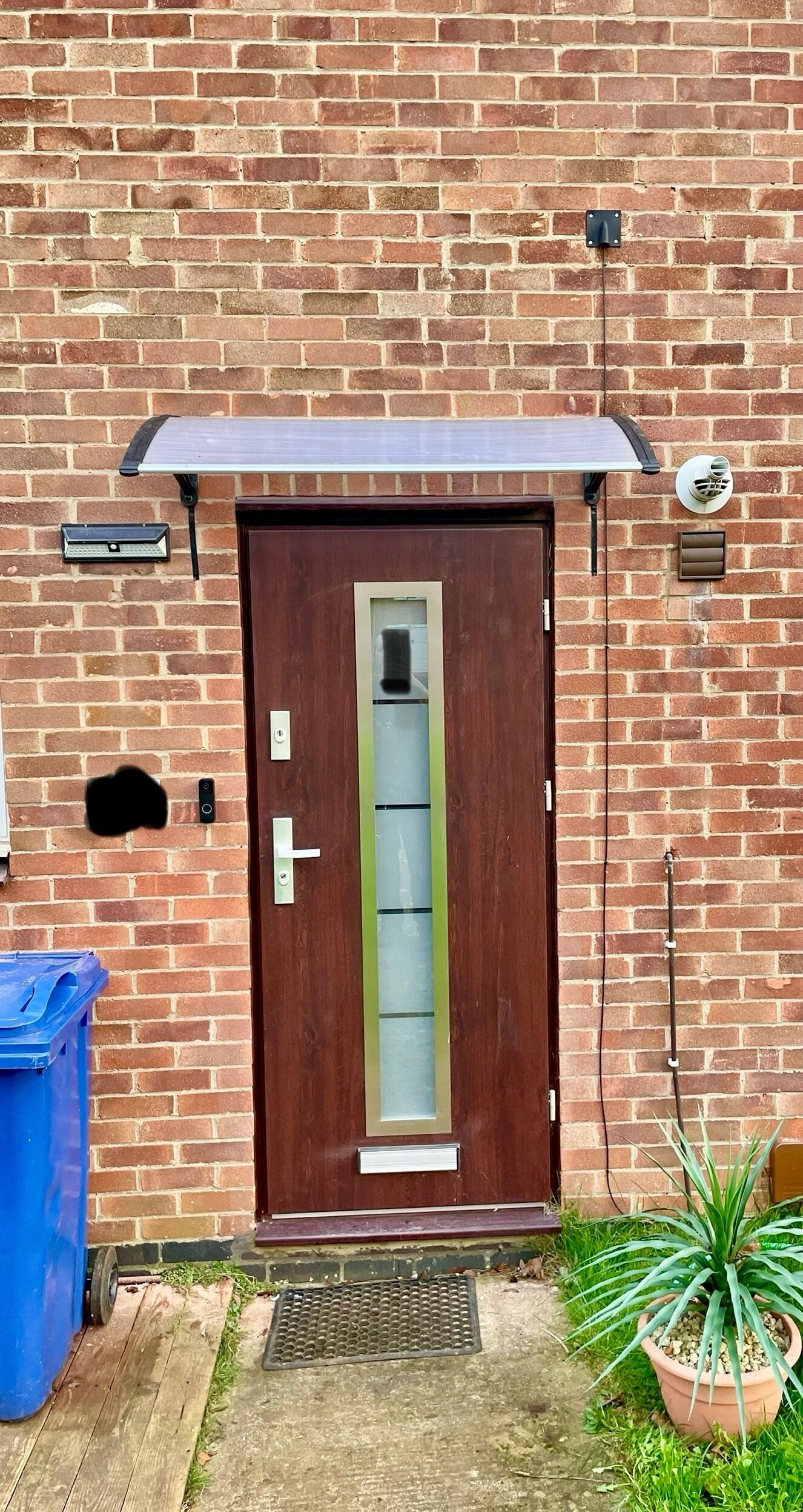 A brick building with a wooden door, a blue trash bin, and a potted plant.