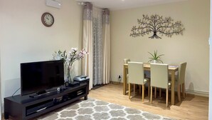 A living room with a flat-screen TV on a black stand, a dining table with four chairs, a wall-mounted tree decoration, and a clock on the wall. - Charming 4-Bed Home, Priv Entrance, Garden, Modern Kitchen, Spacious Living Area (Banbury)