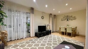 A living room with a TV, a dining table with chairs, a patterned rug, and a decorative tree wall piece. - Charming 4-Bed Home, Priv Entrance, Garden, Modern Kitchen, Spacious Living Area (Banbury)