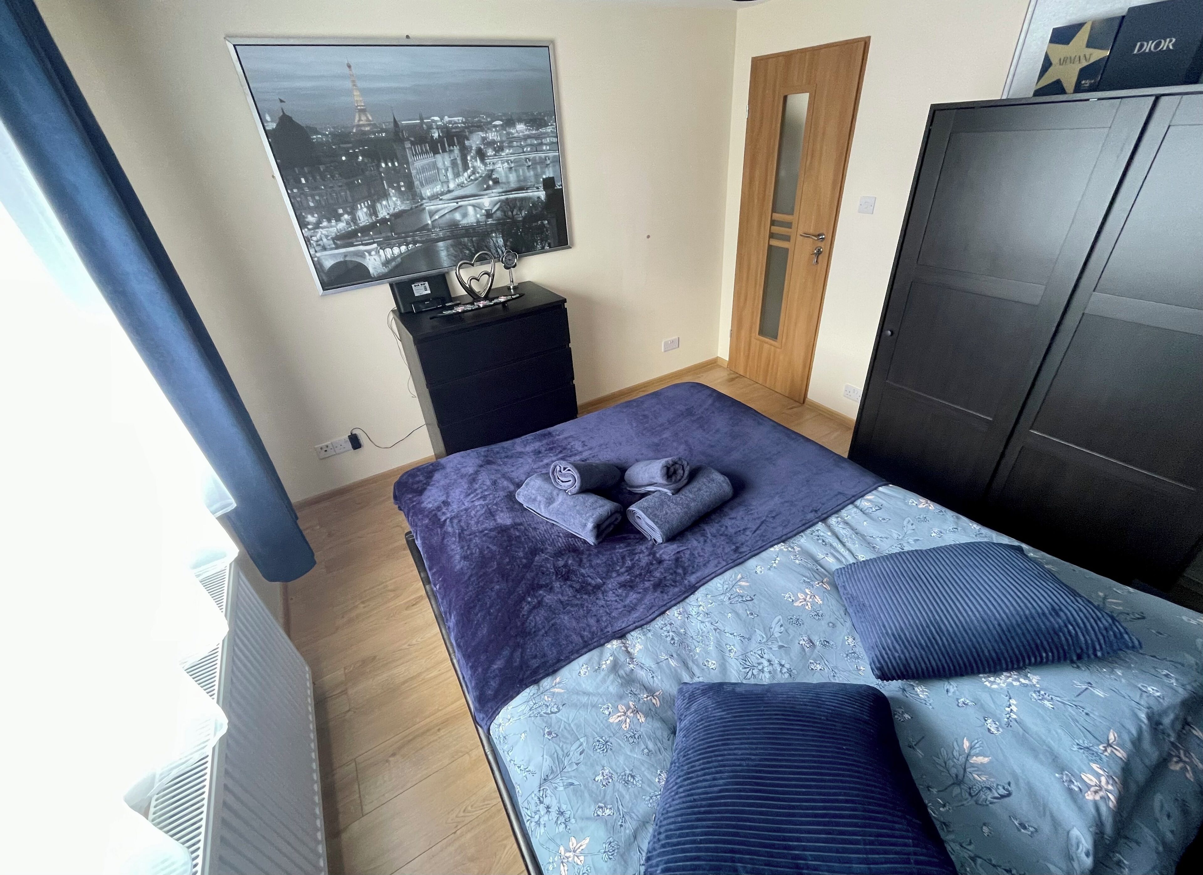 Une chambre à coucher comprenant un lit, une commode, une armoire, un téléviseur fixé au mur et une fenêtre avec des rideaux.