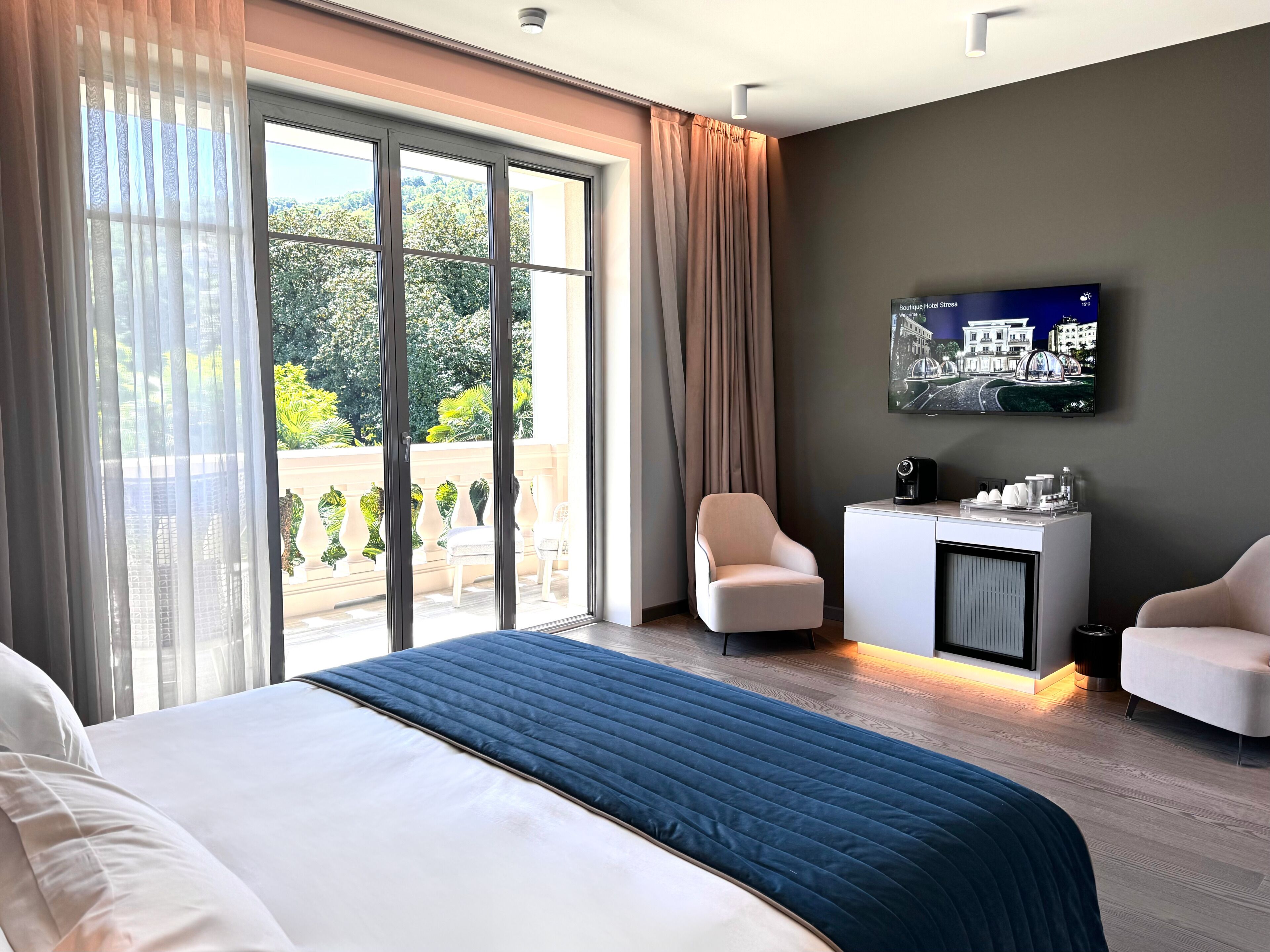 Habitación de hotel con una cama grande, un escritorio con cafetera, un televisor en la pared y un balcón con mobiliario de exterior.