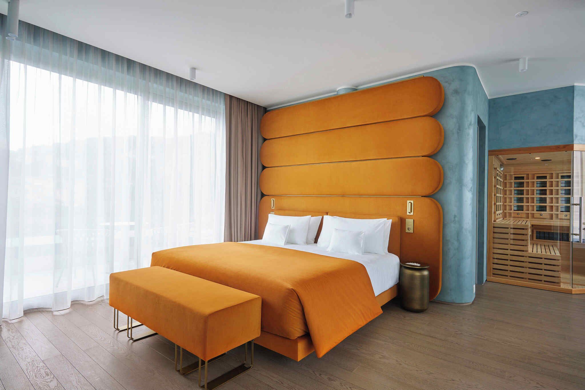 A modern hotel room with a large bed, a bench, and a view of a cityscape through sheer curtains.