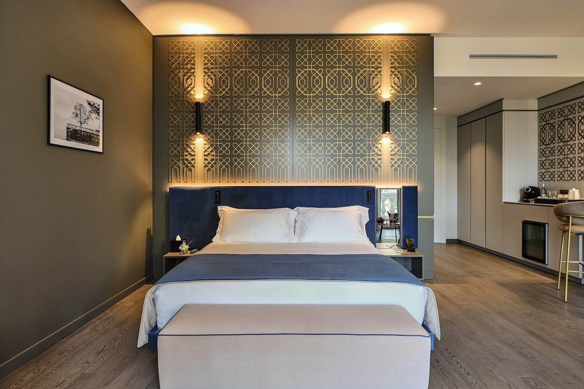 Une chambre d’hôtel moderne dotée d’un grand lit, d’une table de chevet, d’une commode, d’un banc, d’un coin cuisine et d’un panneau décoratif mural.
