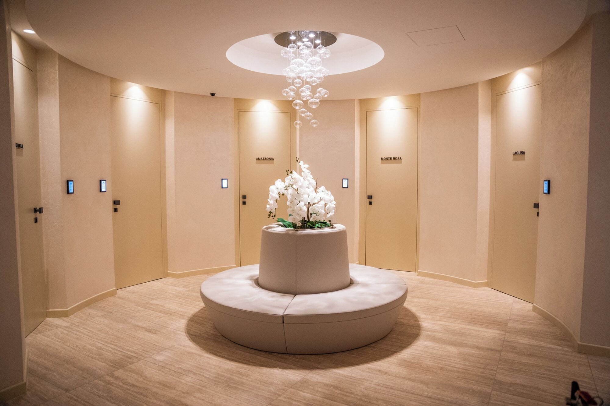 Un hall d’hôtel doté d’un espace salon central circulaire, d’un luminaire décoratif suspendu et de portes portant les inscriptions « AMAZONIE », « MONTE ROSA » et « LAGUNA ».