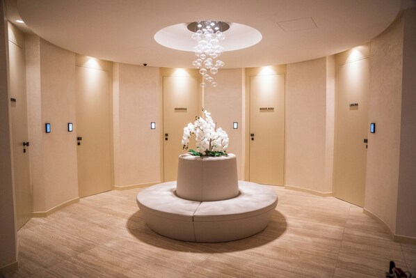 A hotel lobby with a central round seating area, a decorative hanging light fixture, and doors labeled "AMAZONIA," "MONTE ROSA," and "LAGUNA."