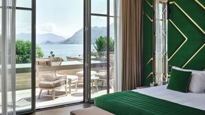 Habitación de hotel con un amplio ventanal corredizo que da a un área exterior con sillas de mimbre y vistas a un lago y montañas.