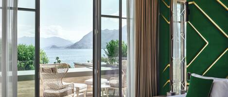Habitación de hotel con un amplio ventanal corredizo que da a un área exterior con sillas de mimbre y vistas a un lago y montañas.