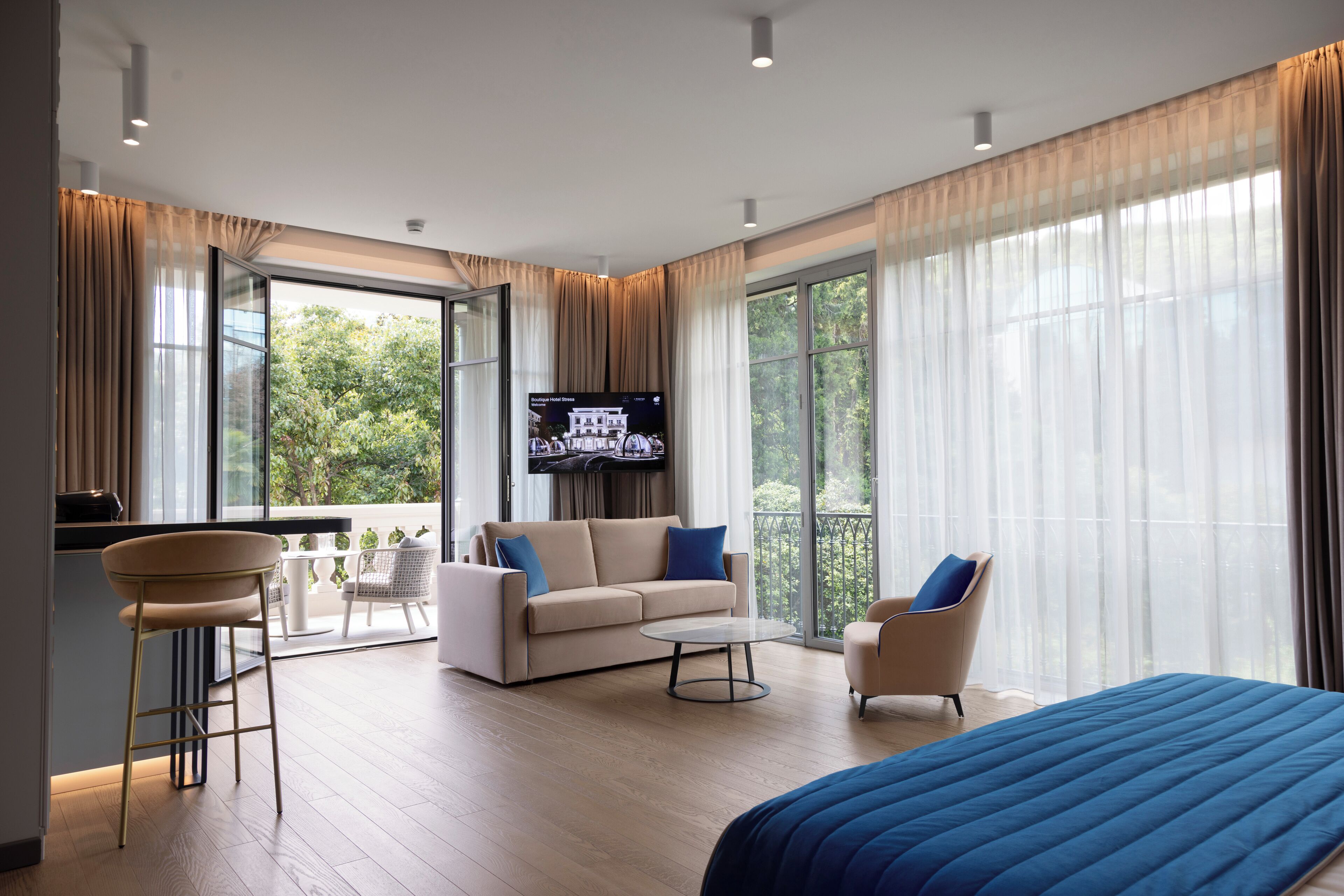 A modern hotel room with a large bed, a sofa, a TV, and a balcony with a view of greenery.