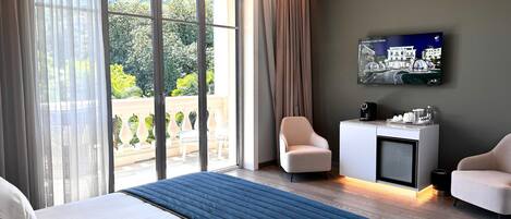 Habitación de hotel con una cama grande, un escritorio con cafetera, un televisor en la pared y un balcón con mobiliario exterior.