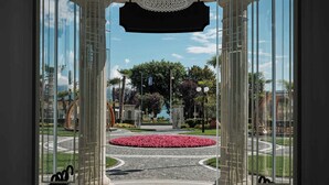 Vista a través de un ventanal que da a un patio con un parterre central, una fuente y cielo despejado.