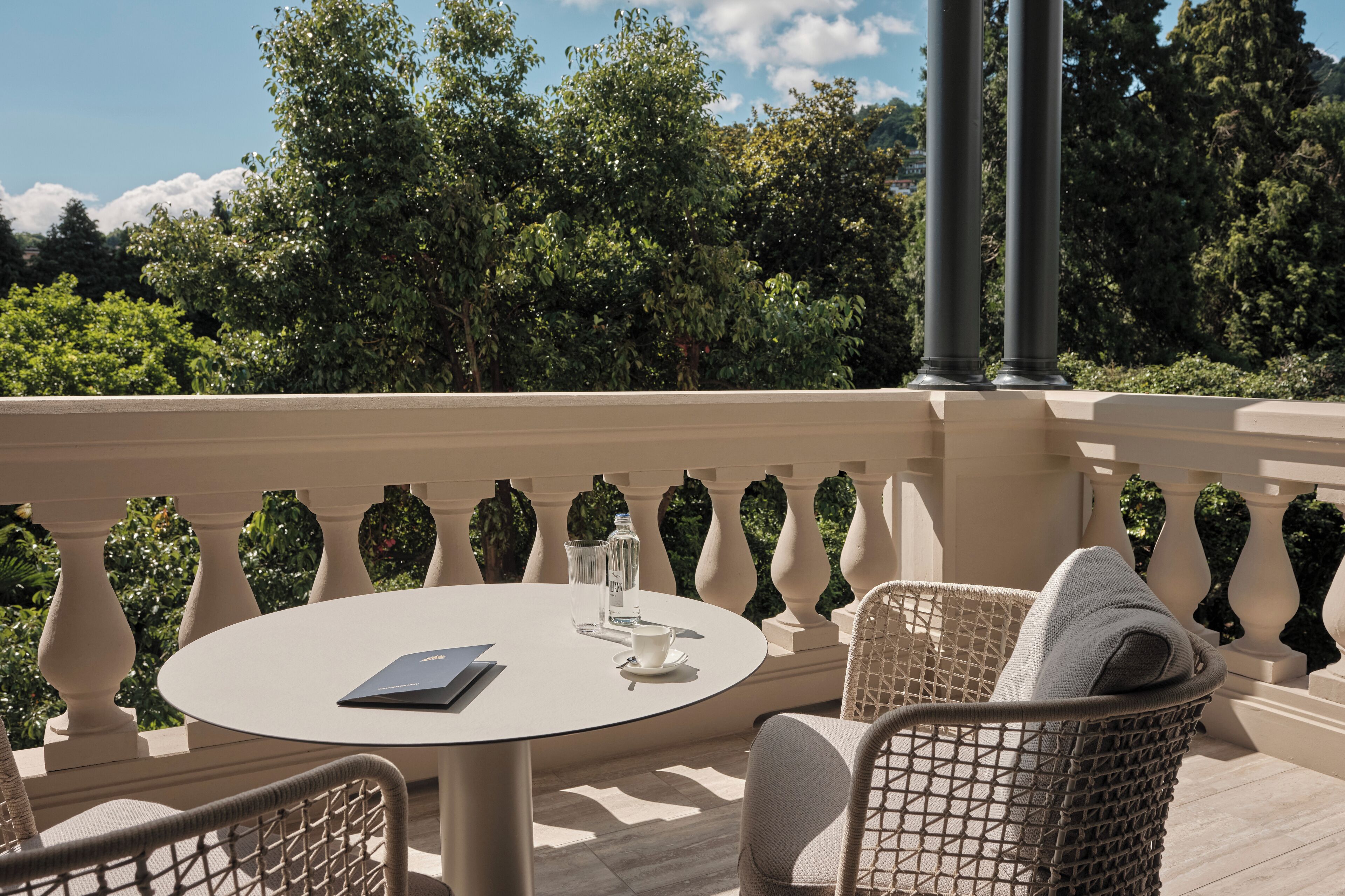 A round table with a book, a glass, and a cup on a balcony with wicker chairs and a balustrade.