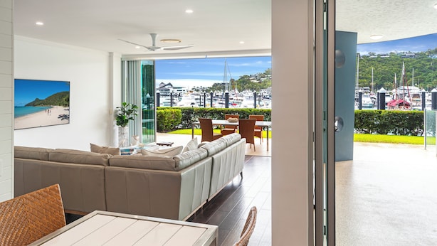 A modern living room with a sofa, dining table, and chairs. A large window offers a view of a marina with boats. - Pavillions 6, Pavilions 6 on Hamilton Island (Hamilton Island)