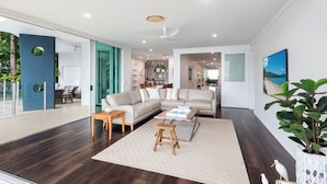 A modern living room with a sectional sofa, a coffee table, and a small stool. There is a large window with a view of the outdoors. - Pavillions 6, Pavilions 6 on Hamilton Island (Hamilton Island)