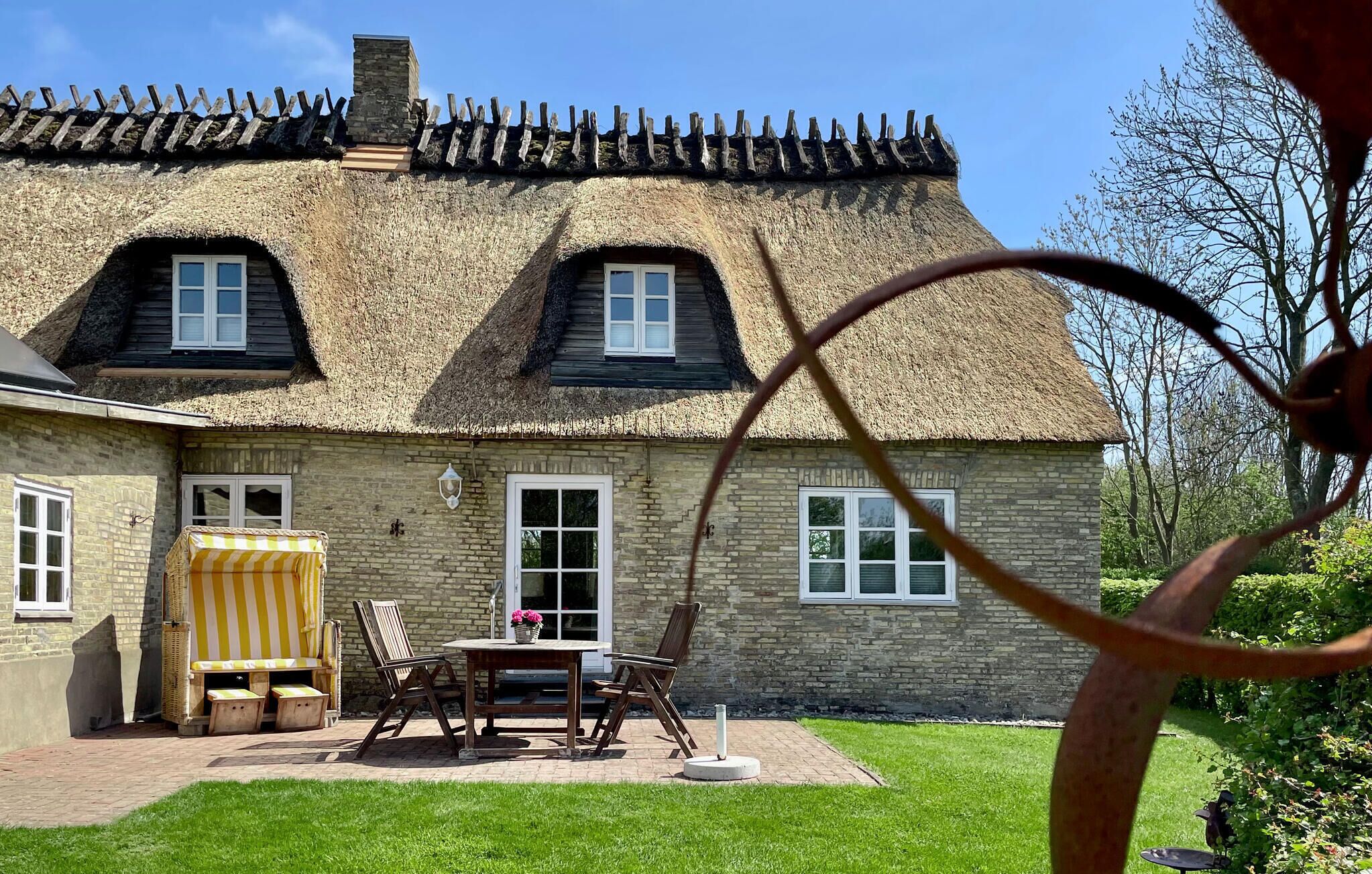 Une maison traditionnelle en pierre avec un toit de chaume, une terrasse en briques avec des meubles d’extérieur et une pelouse bien entretenue.