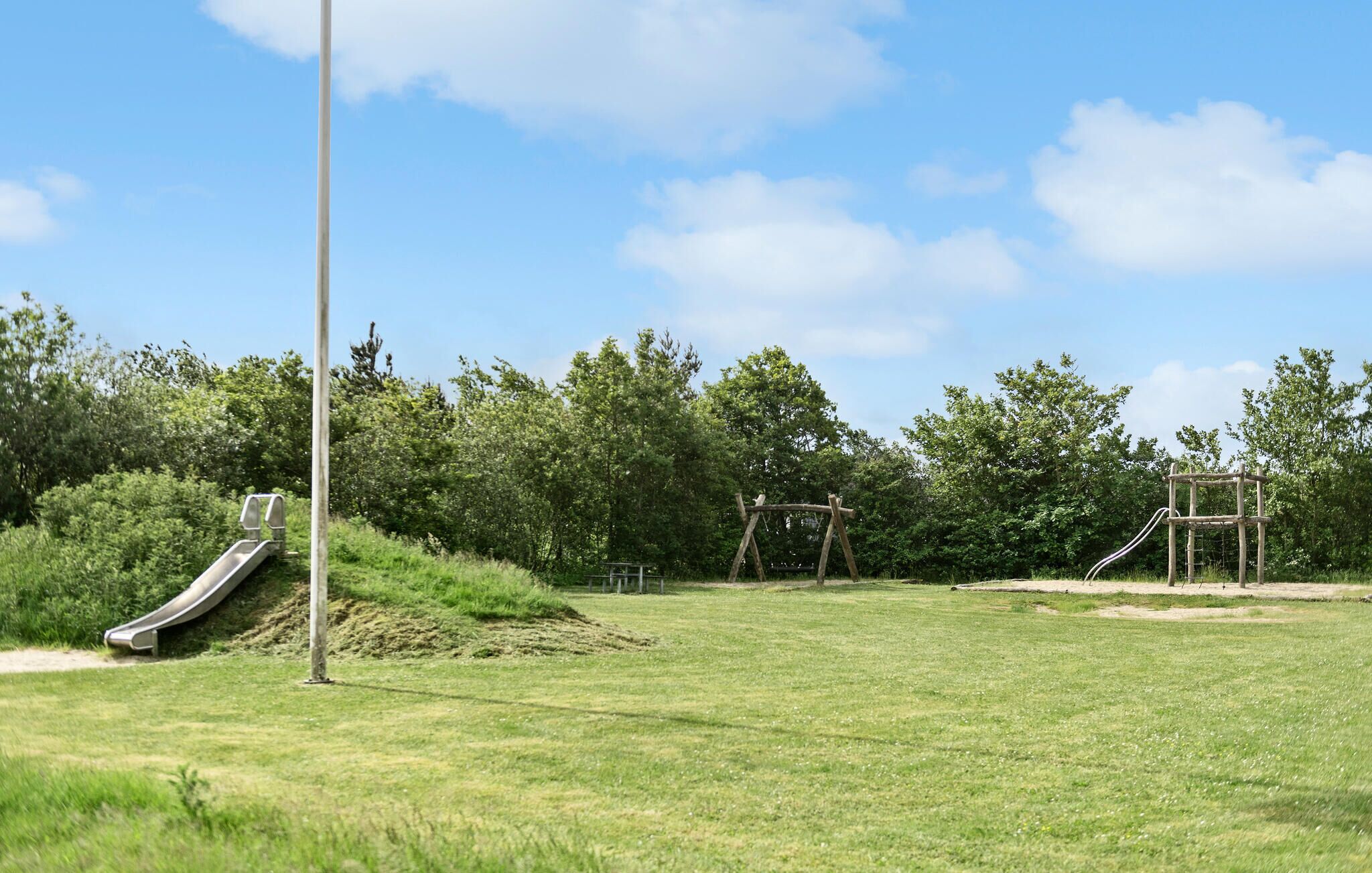 Une aire de jeux avec un toboggan et des balançoires, située dans un espace verdoyant avec des arbres en arrière-plan.