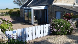 Une maison en pierre dotée d’un espace extérieur couvert avec des sièges, d’une allée en gravier et d’une clôture blanche sur laquelle est affiché un numéro.