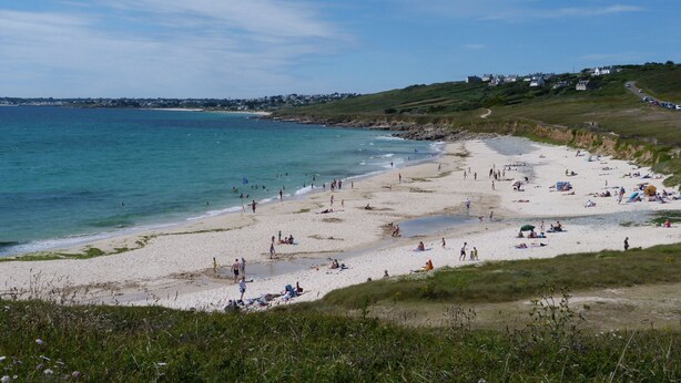 Une plage où des gens se prélassent au soleil et se baignent, avec des maisons en arrière-plan et un ciel dégagé.