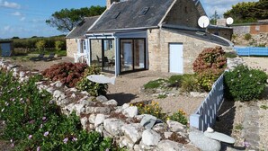 Une maison en pierre avec un toit en pente, une porte d’entrée en verre et un patio aménagé avec du mobilier d’extérieur.