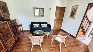 Ein Zimmer mit einem dunkelblauen Sofa, zwei Korbstühlen, einem Glastisch und einem Holzkleiderschrank sowie einer Holztür.