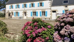 Un bâtiment à deux étages, doté d’une façade en pierre et de volets bleus, entouré d’une végétation luxuriante et de fleurs d’hortensia.