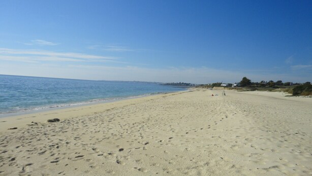 Une vaste plage de sable fin, parsemée d’empreintes menant à l’eau, un ciel dégagé et des bâtiments qui se dessinent à l’horizon.