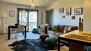 A modern living room with a sofa, a dining table, and a kitchen area with a cutting board and utensils. - Higgi Drive 5 - Mt Hotham (Hotham Heights)