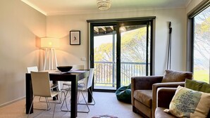 A cozy living room with a dining area, a sofa, and a view of a balcony with trees. - Higgi Drive 5 - Mt Hotham (Hotham Heights)