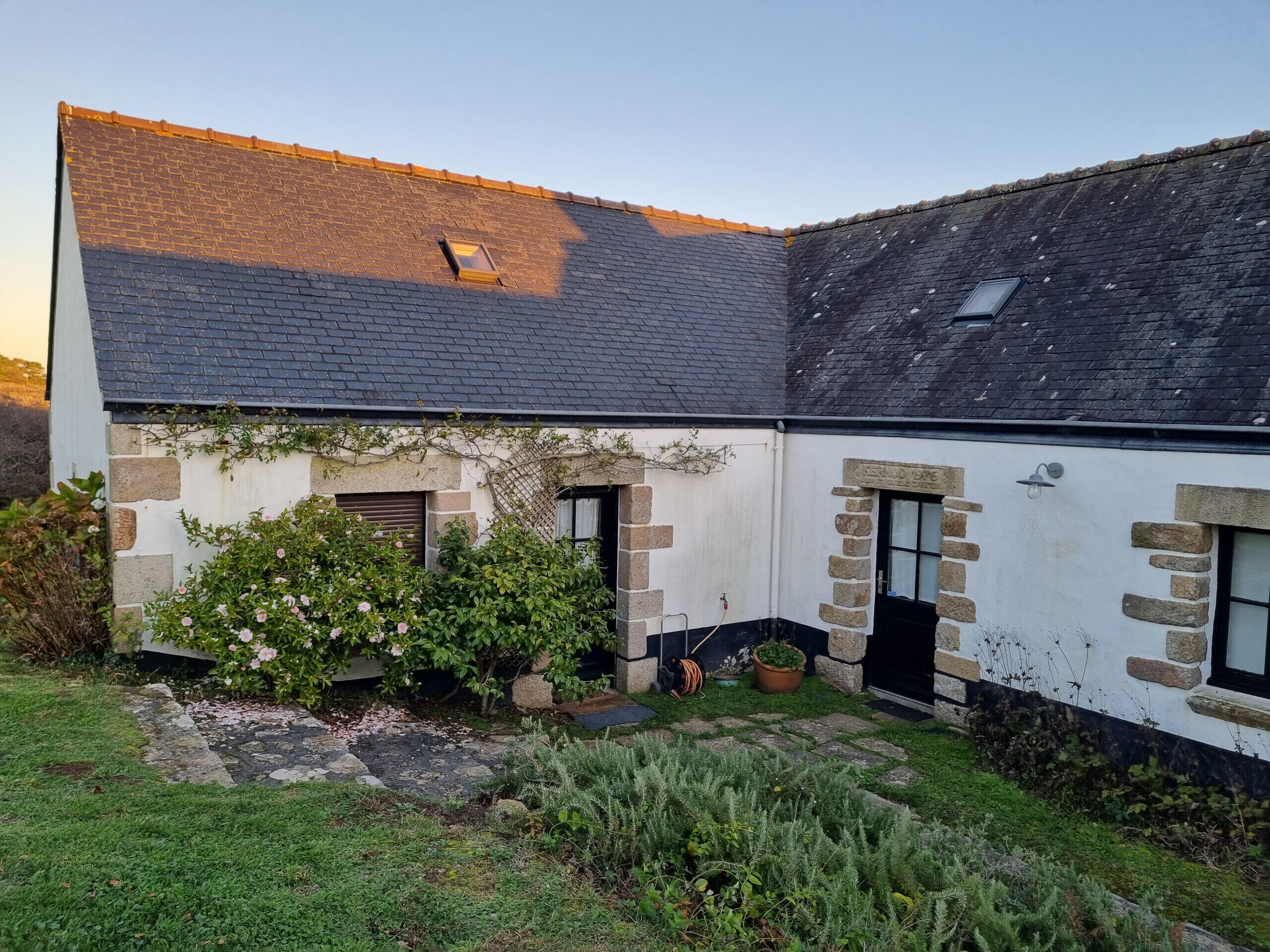 A two-story house with a sloped roof, stone walls, and a small garden area with flowering plants.