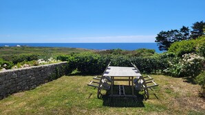 A wooden table and chairs set on a grassy lawn with a stone wall and flowering plants, overlooking a coastal landscape.