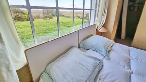 A room with a large window offering a view of a green field. There are two beds with white bedding and a lamp on a bedside table.