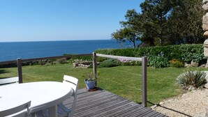 A wooden deck with a white table and chairs, overlooking a garden with a view of the sea.
