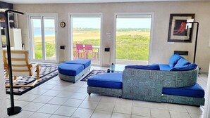A spacious living room with a large sofa, a chair, and a small table. There are large windows offering a view of the beach and greenery. - 622-Ker An Enez,sea view, the beach at the end of the garden (Esquibien)