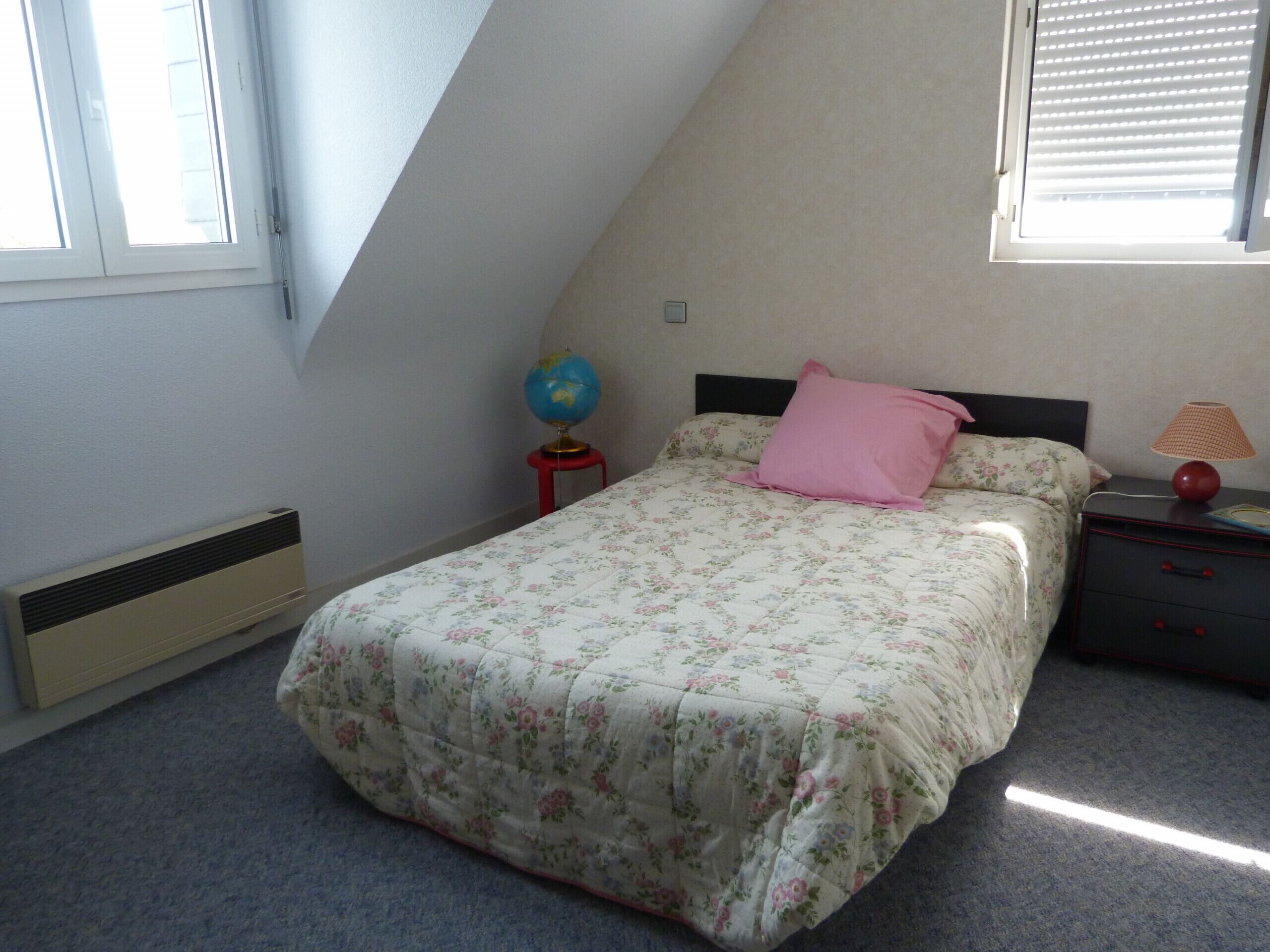 A bedroom with a sloped ceiling, a bed with floral bedding, a globe on a small stool, a pink pillow, a nightstand with a lamp, and a radiator.