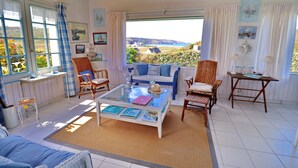 Una habitación acogedora con un gran ventanal, sillas de mimbre, una mesa de centro con libros y vista al mar.