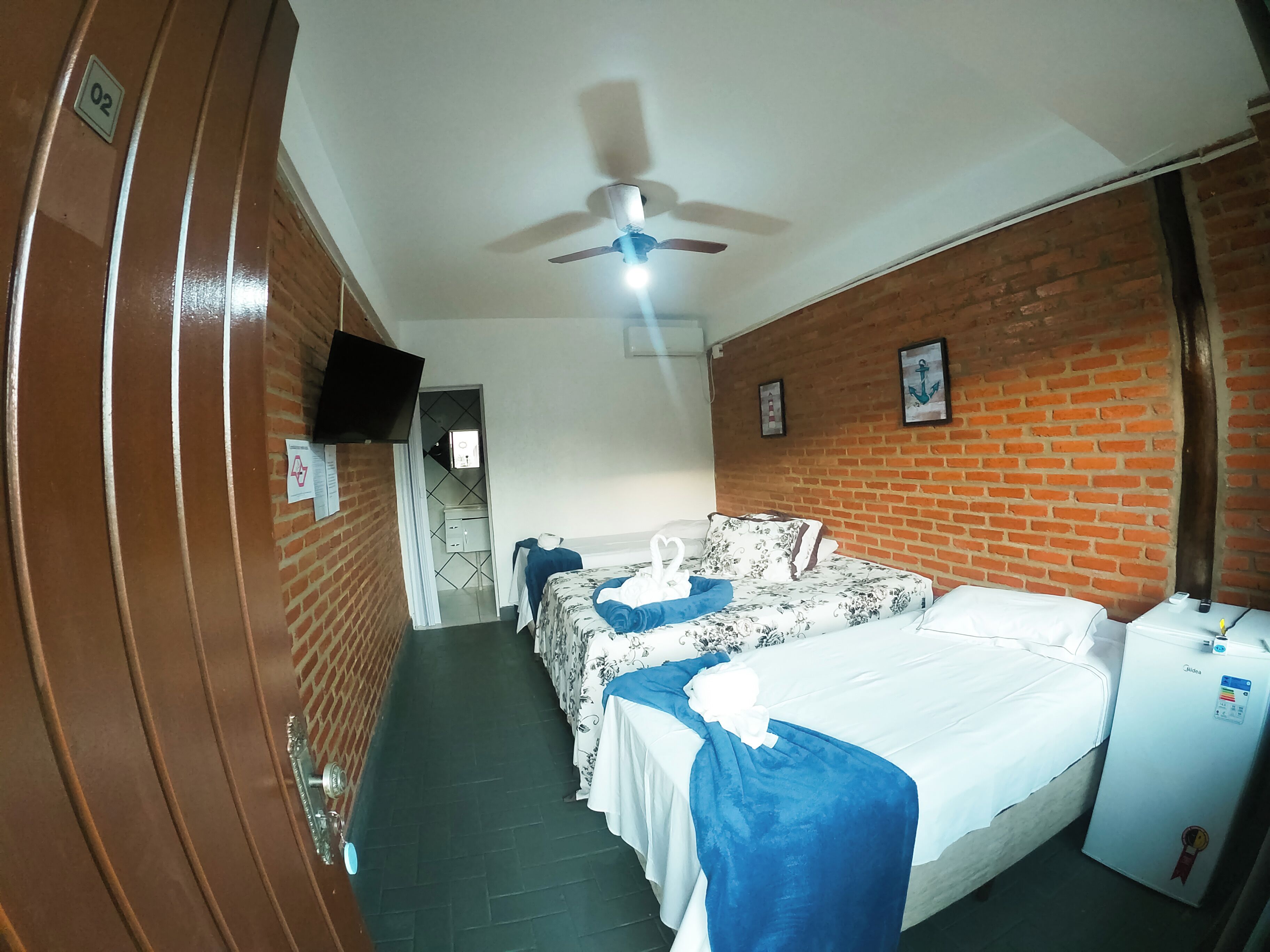 A hotel room with a bed, a ceiling fan, a brick wall, and a small refrigerator.