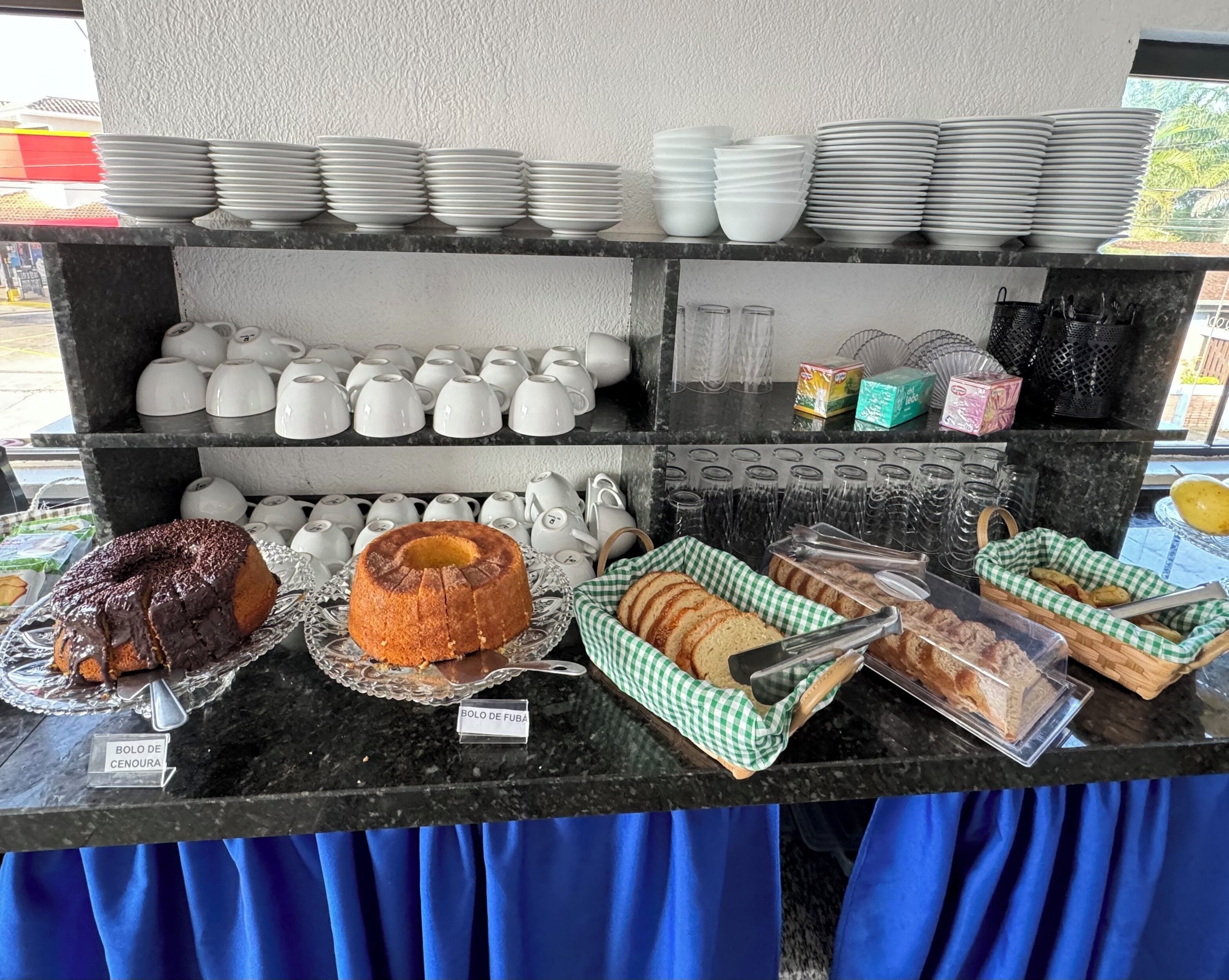 A buffet counter with various food items and utensils, including a chocolate-covered cake, a bundt cake, and several trays of bread.