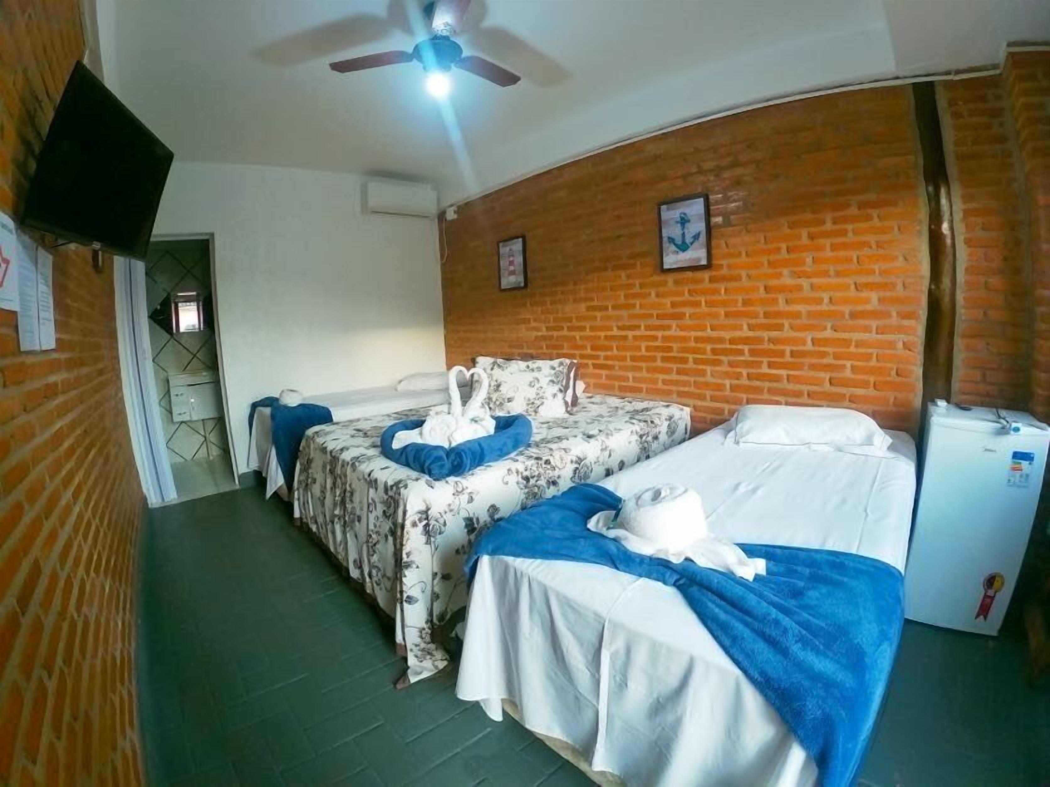 Habitación de hotel con dos camas, ventilador de techo, refrigerador y pared de ladrillo.