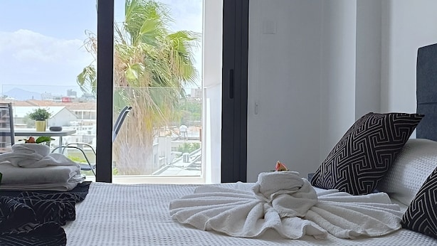 A hotel room with a large bed, a view of palm trees and buildings, and a balcony. - Larvella Estia Luxury Suites (Larnaca)