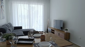 A modern living room with a grey sofa, a wooden dining table set for two, and a flat-screen TV on a wooden stand. - Larvella Estia Luxury Suites (Larnaca)