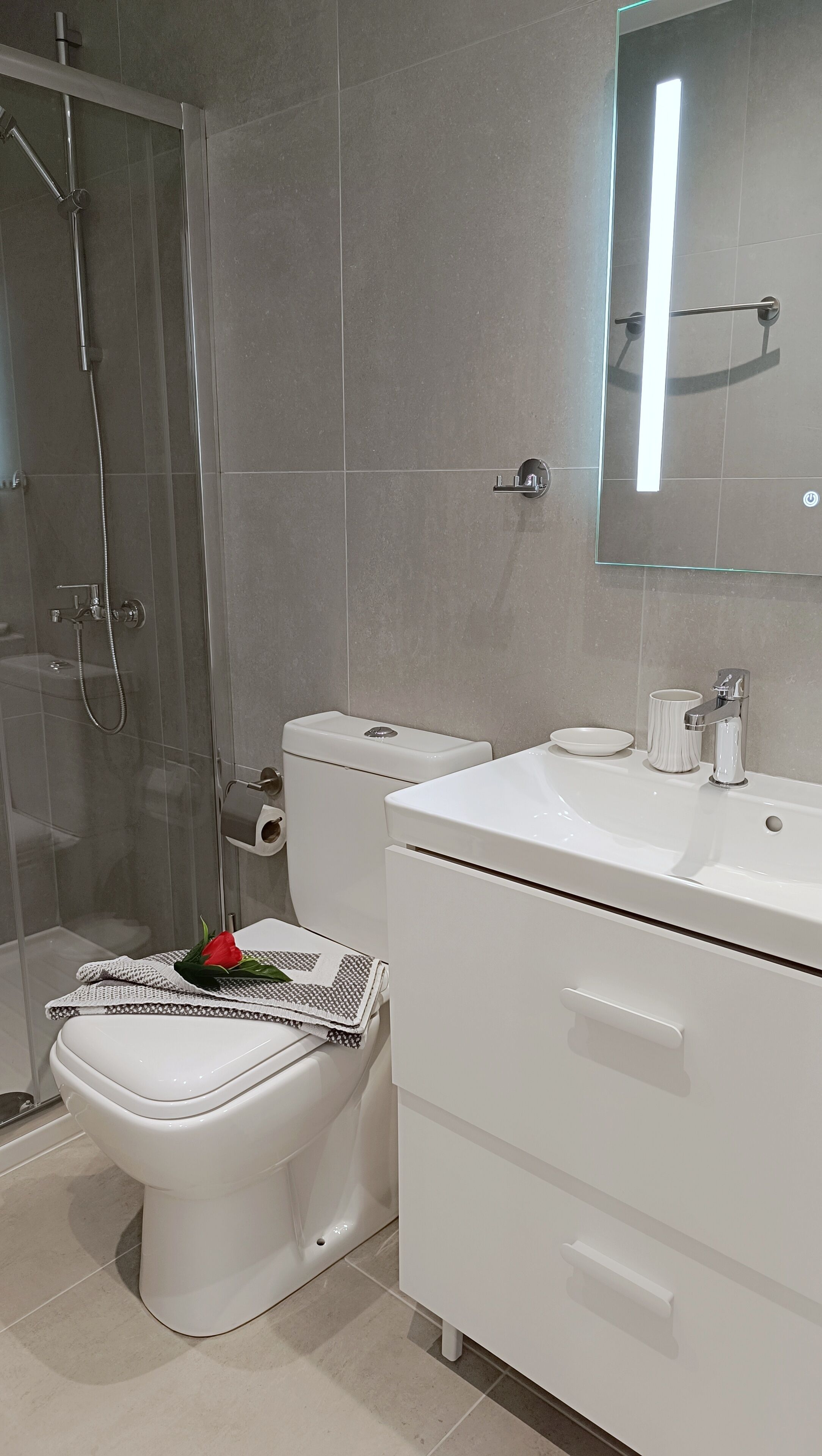 Un baño moderno con cabina de ducha de vidrio, un inodoro con un tapete decorativo y un tocador blanco con espejo.