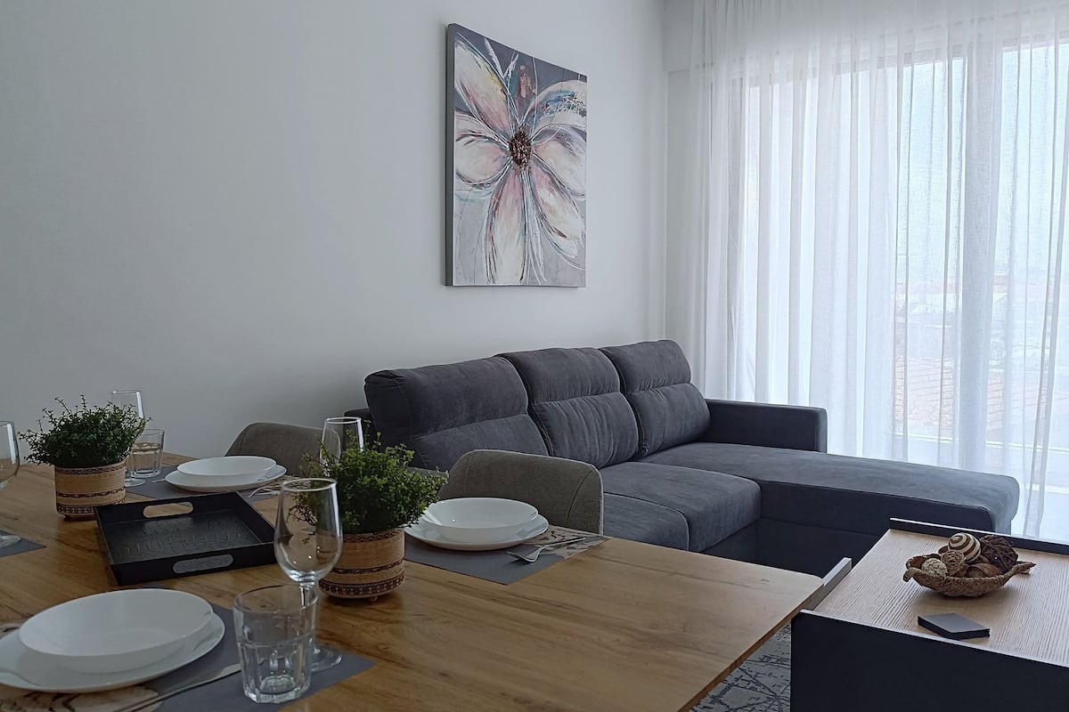 Una sala moderna con comedor. Incluye un sofá gris, una mesa de comedor de madera con platos y vasos, y un cuadro abstracto grande en la pared.
