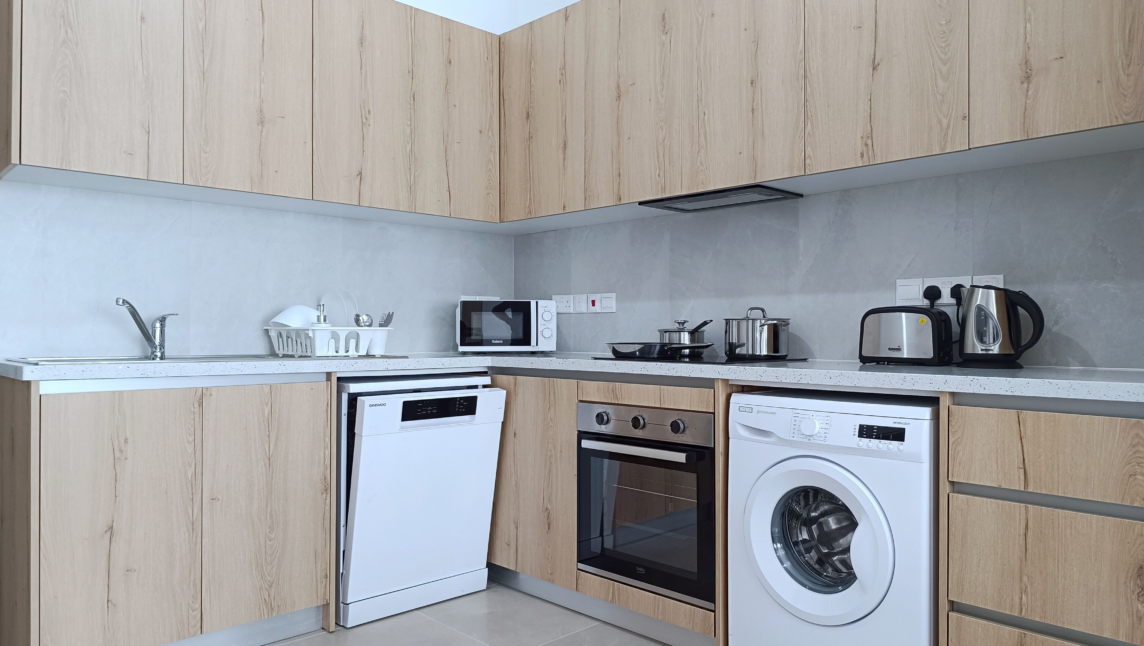 Eine kleine Küche mit Holzausstattung, einem Geschirrspüler, einer Waschmaschine, einer Mikrowelle, einem Toaster und einem Wasserkocher.