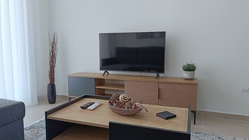 Egy modern nappali, melyben egy síkképernyős televízió áll egy fa állványon, egy kávézóasztalon távirányítók és dísztárgyak találhatók, egy oldalasztalon egy virágos növény, és egy magas váza, melyben ágak vannak.