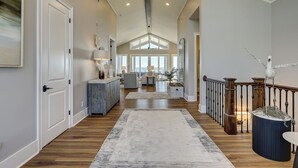 A spacious hallway with wooden flooring, a central rug, and a view to a living area with a large window overlooking the ocean. - 11 Sea Hawk Lane - Oceanfront Oasis: Stylish Coastal Living & Endless Views (Hilton Head Island)