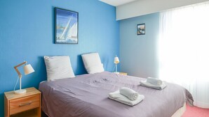 Un lit bien fait, recouvert d’une couvre-lit violette et agrémenté d’oreillers blancs, une table de chevet en bois avec une lampe, et un tableau encadré accroché au mur.
