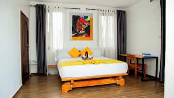 A bedroom with a wooden bed, white bedding, and two orange pillows. There is a wooden desk and chair, a radiator, and a painting on the wall.